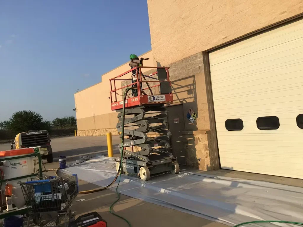 A worker on a red scissor lift uses a hose or sprayer to work on the exterior wall beside a large roll-up loading dock door, with plastic sheeting spread on the ground and equipment staged nearby in the parking area.