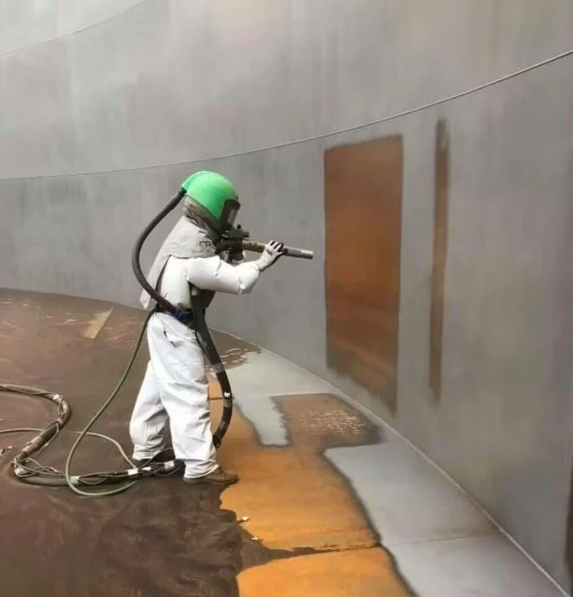 A worker in a white blast suit with a green helmet uses a hose-fed blasting nozzle to clean or strip coating from the curved interior wall of a large tank, revealing rust-colored patches and a lighter cleaned area on the floor and wall.