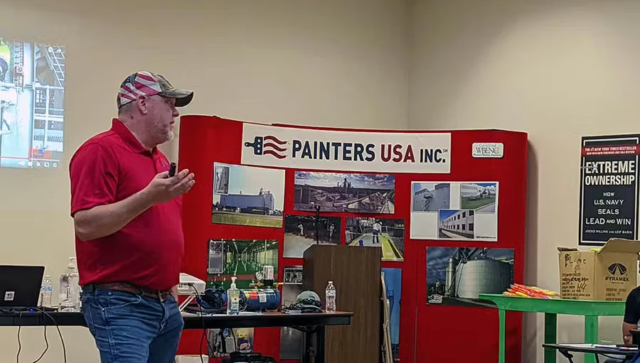A man in a red polo and jeans speaks to a group in a training room, standing beside a red display backdrop labeled “PAINTERS USA INC.” with photos of industrial job sites; tables with safety gear and materials sit in front.