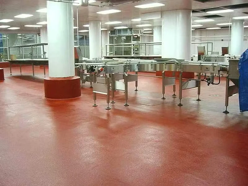 Interior of a commercial food-processing area with stainless-steel conveyor equipment and large white columns, featuring a red textured epoxy-style floor under bright overhead lighting.