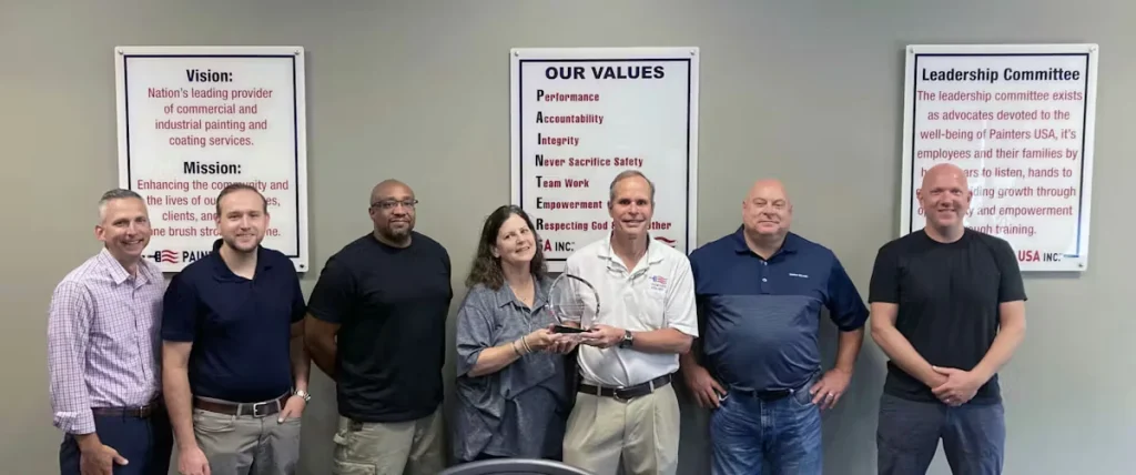 Group photo of seven adults standing indoors against a gray wall with three framed posters (“Vision/Mission,” “Our Values,” and “Leadership Committee”); two people in the center hold a clear glass award while the others pose on either side.