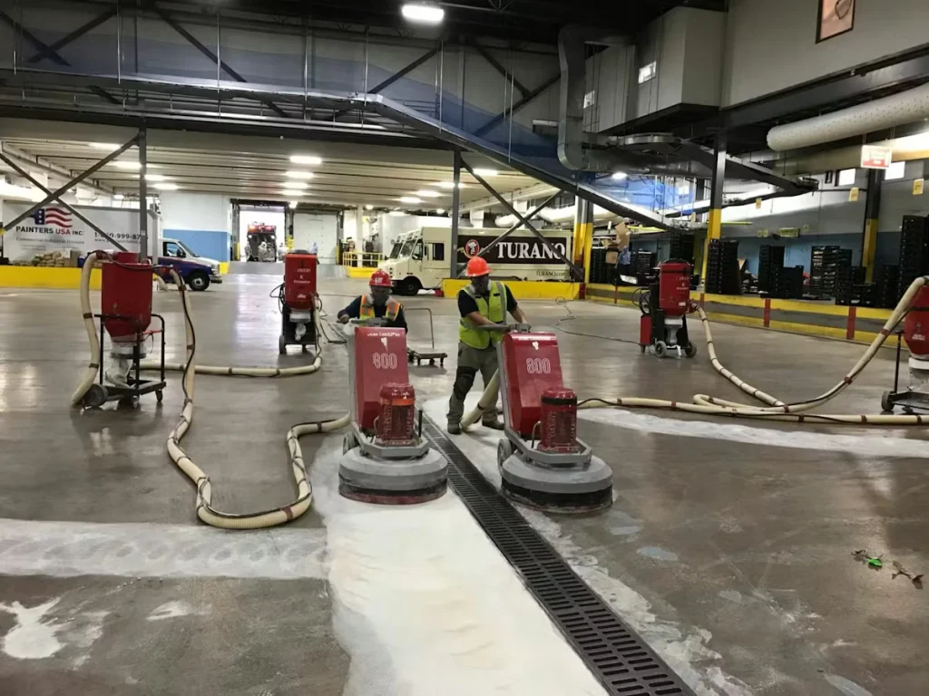 Inside a large industrial facility, two workers in safety vests and hard hats operate red floor-grinding/polishing machines connected to vacuum hoses, with multiple other machines and hoses spread across the concrete floor.