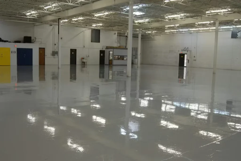 Wide view of a large, mostly empty warehouse with a high white ceiling and columns, where a smooth gray floor coating reflects the overhead lights and nearby doors and walls.