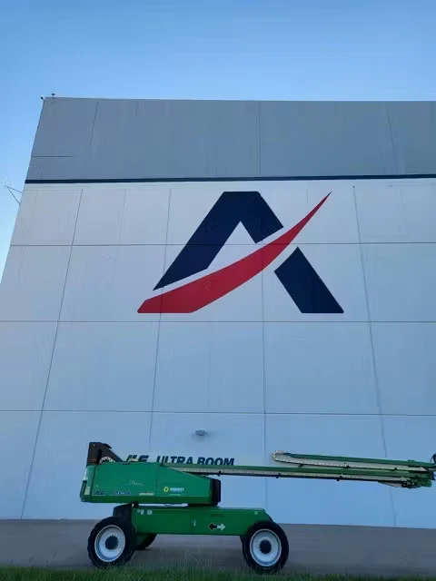 Tall white industrial building facade with a large dark blue stylized “A” and a red swoosh logo centered on the wall; a green boom lift is parked along the bottom edge under a clear blue sky.