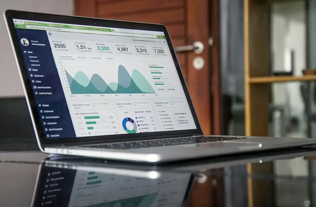 An open laptop on a glossy table displays an analytics dashboard with charts, graphs, and KPI numbers, with an office doorway and shelving blurred in the background.