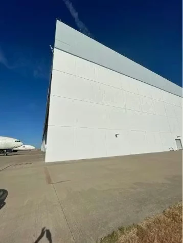 A large white industrial building with tall, smooth exterior wall panels sits beside a wide concrete apron under a clear blue sky, with part of an airplane visible at the left edge.