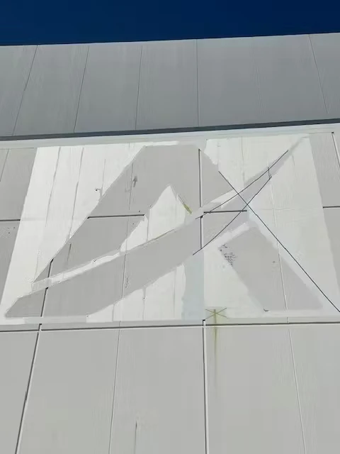 Close-up of a white industrial wall where a large logo shape is being painted; taped guide lines and a diagonal rope or cable cross the panel seams under a deep blue sky.