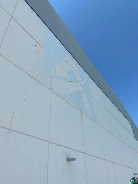 Low-angle view of a tall white panelized building facade under a clear blue sky, showing faint outlines of a large logo sketched high on the wall and a small wall-mounted light fixture below.