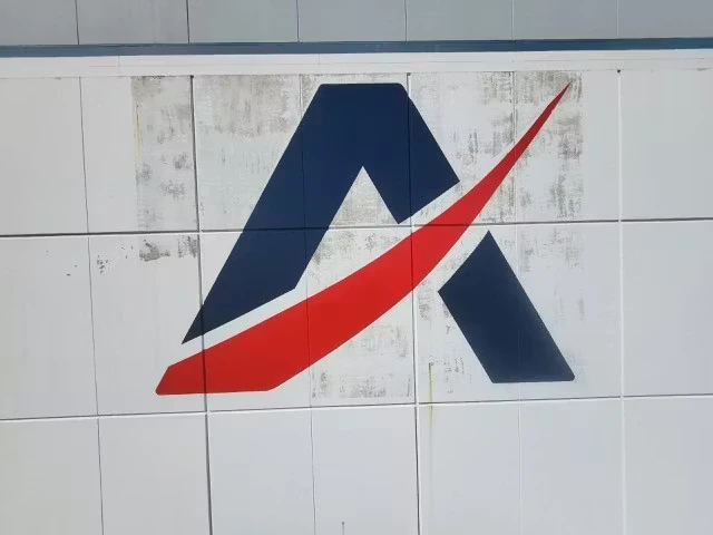 Completed logo painted on a white, grid-paneled exterior wall: a dark blue angular “A” shape with a red swoosh sweeping diagonally across it, with slight weathering marks visible around the panels.
