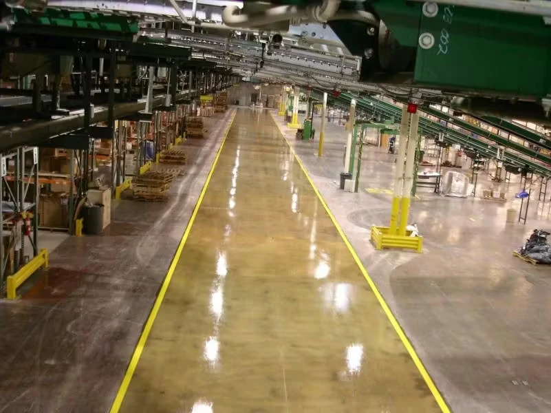 High-angle view down a long factory aisle with a glossy floor marked by bright yellow lines, surrounded by conveyors, machinery, and stacked pallets under industrial lighting.