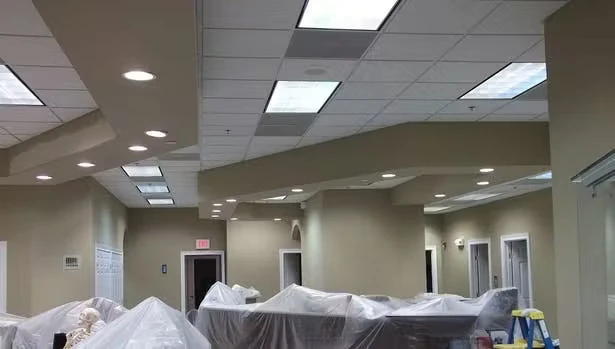Office interior under renovation with beige walls, a drop ceiling with rectangular fluorescent light panels, and furniture covered in plastic sheeting, with a ladder visible on the right.