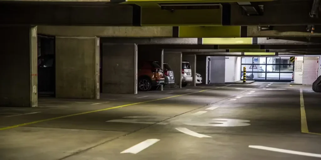 Dimly lit underground parking garage with painted directional arrows and lane lines on the concrete floor, several cars parked in bays along the left, and daylight visible at the far exit.