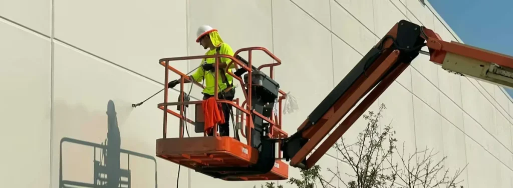 Worker in a white hard hat and neon safety shirt stands in an orange boom lift basket while pressure-washing a large white tilt-up concrete wall; the lift arm extends from the right with tree branches visible below.