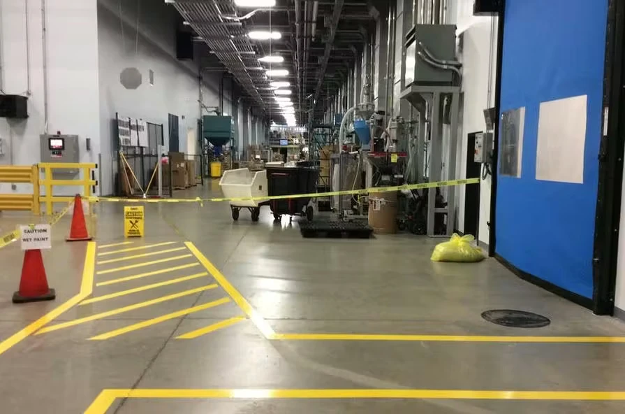 Industrial facility corridor with freshly painted yellow floor striping and diagonal hatch markings near a taped-off work area; orange cones and caution signs sit on the polished concrete floor beside machinery and equipment in the distance.