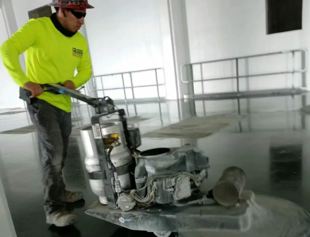 Worker in a neon-yellow shirt and hard hat operates a floor polishing machine on a glossy concrete surface inside an industrial facility, with wall-mounted rails in the background.