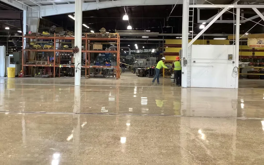 Wide view of an industrial shop with a highly polished concrete floor reflecting overhead lights; shelving and equipment line the back, and two workers in high-visibility shirts stand near an open bay.