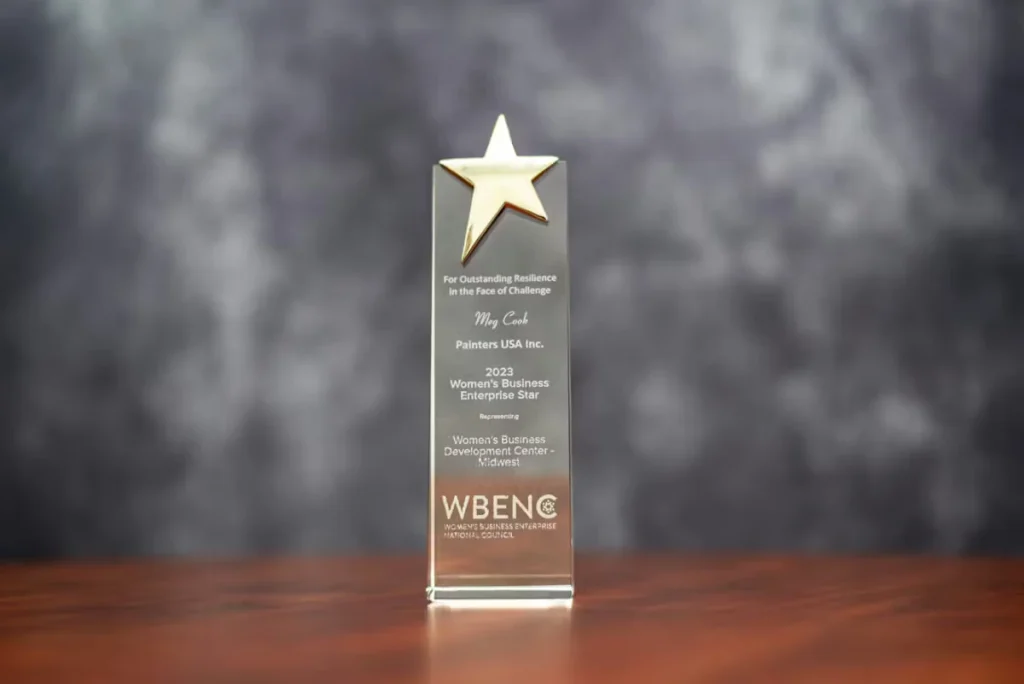Tall, rectangular glass award topped with a gold star, engraved with recognition text including “Meg Cook,” “Painters USA Inc.,” and “2023 Women’s Business Enterprise Star,” with WBENC logo near the bottom, sitting on a wooden surface against a blurred gray backdrop.