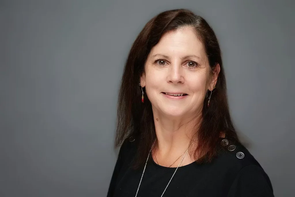 Studio-style headshot of Painters USA President and Board Chair Meg Cook with shoulder-length dark brown hair, smiling slightly, wearing a black top and small drop earrings, photographed against a smooth gray background.
