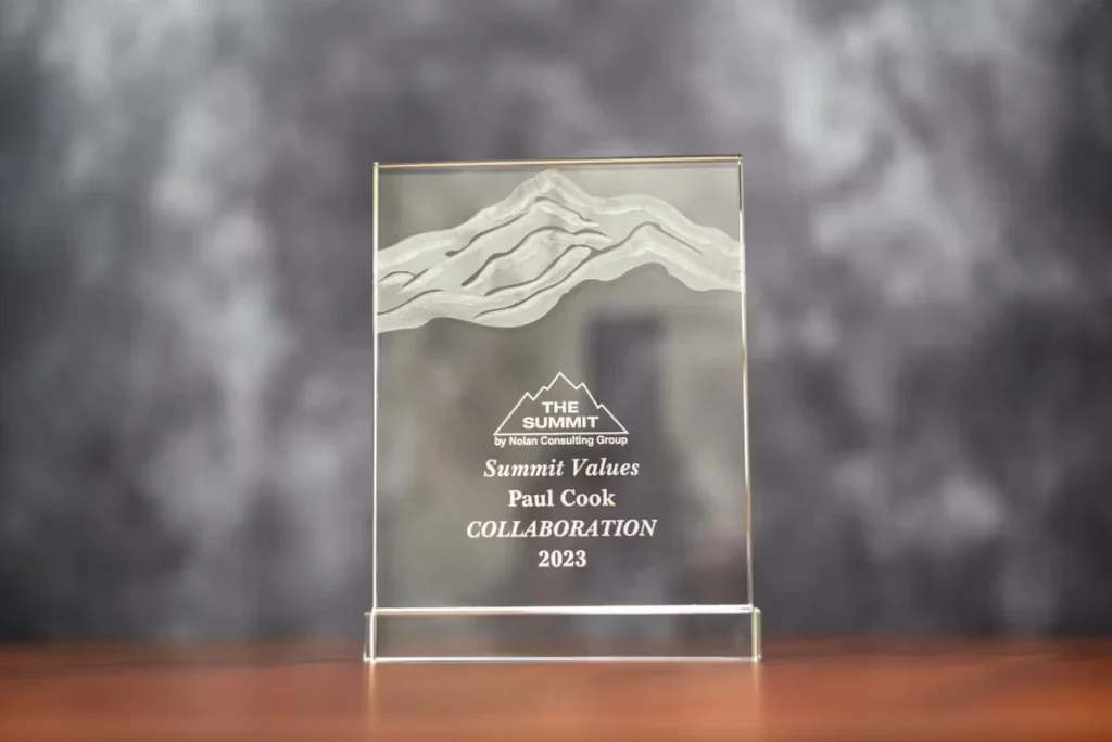 Clear glass award with an etched mountain design at the top, showing the logo “THE SUMMIT by Nolan Consulting Group” and the text “Summit Values Paul Cook COLLABORATION 2023,” sitting on a wooden surface against a gray blurred background.