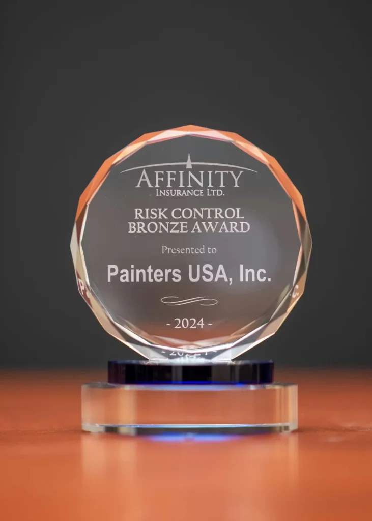 A clear, faceted glass award on a blue-and-clear base sits on a wooden surface, engraved “Affinity Insurance Ltd. Risk Control Bronze Award, Presented to Painters USA, Inc., 2024,” against a dark blurred background.