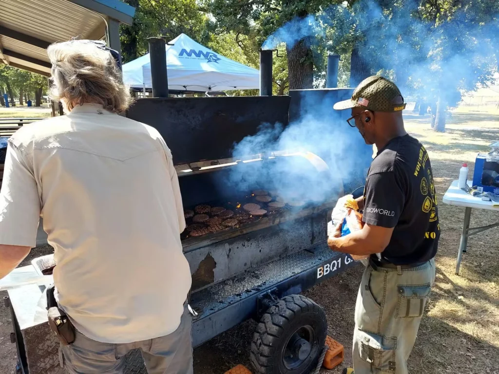 Two people stand at a large outdoor barbecue smoker as thick smoke rises; burgers cook on the grill while a man in a cap and dark T-shirt tends the smoker, with picnic tables and a canopy tent in a park setting behind them.
