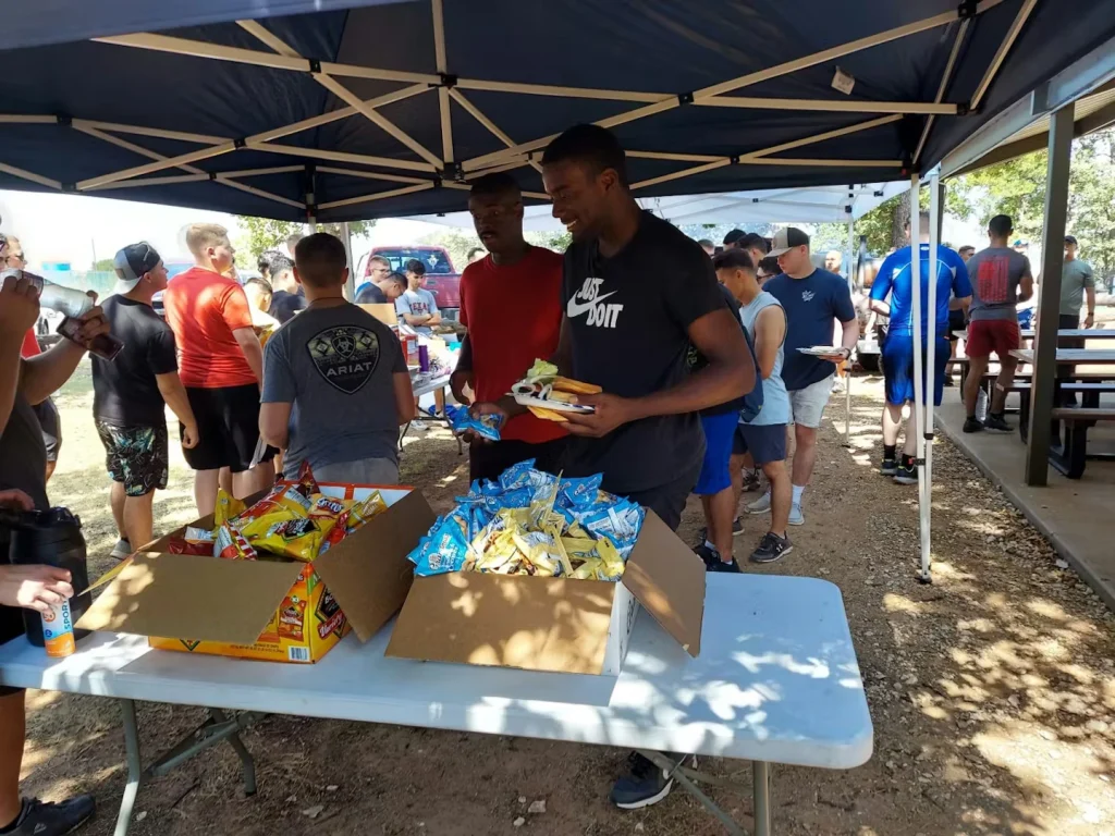 Under a pop-up canopy at an outdoor gathering, people line up for food while a man holds a plate and reaches toward a table with open boxes of assorted snack chips and drinks.