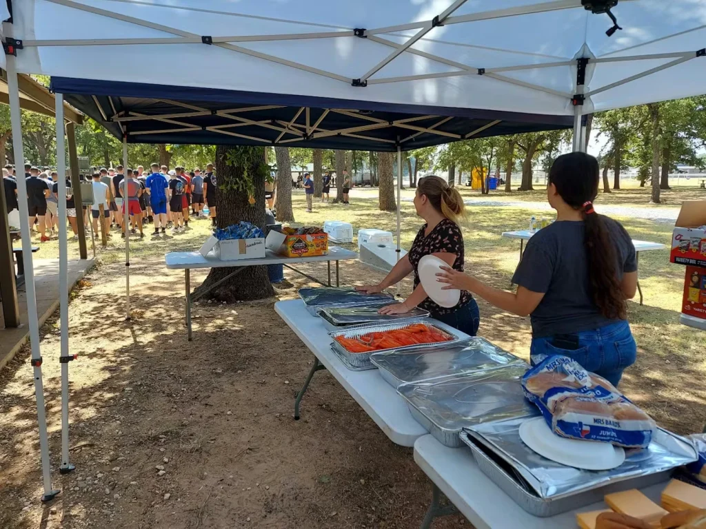 Two women stand behind folding tables under a canopy at an outdoor event, with covered catering trays, buns, and snacks set out while a group of people gathers in the background near trees.