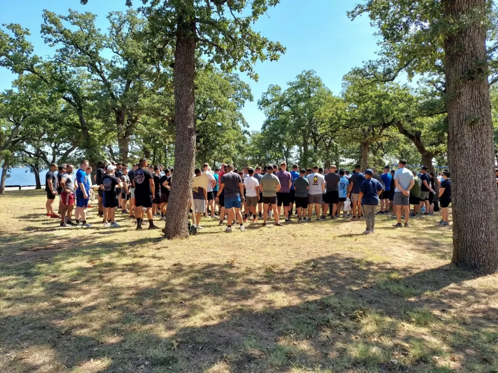 A large group of people stands in a loose cluster under tall trees in a sunny park, facing inward as if listening to someone, with open grass and a glimpse of water in the distance.