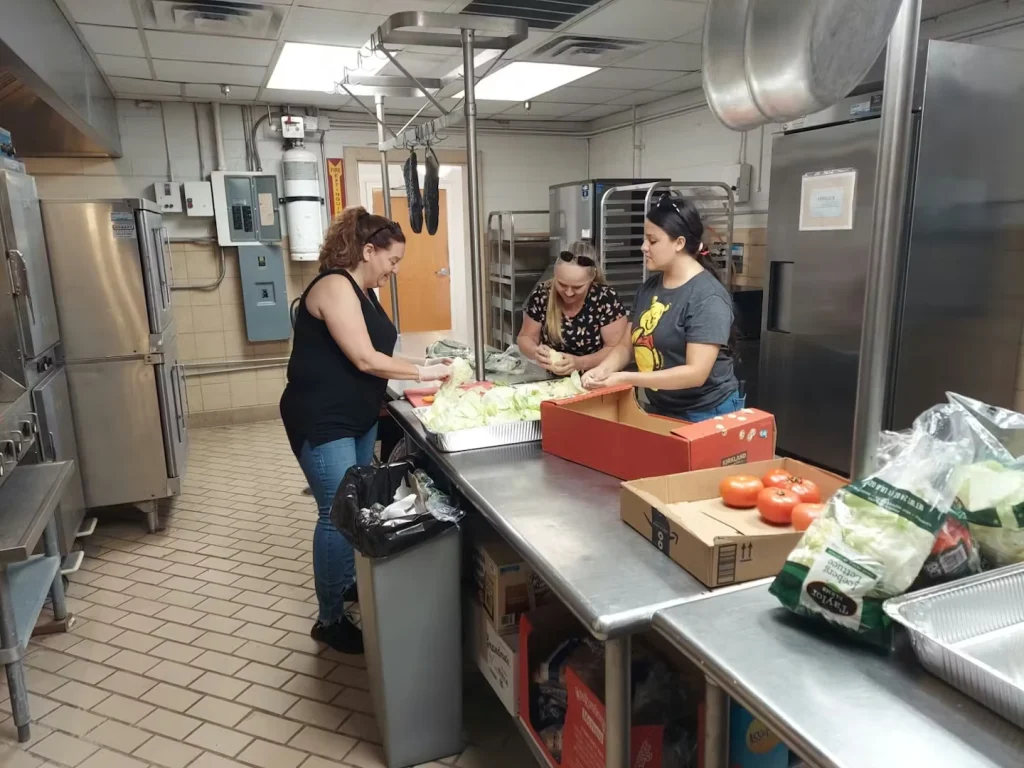 Three people prepare food at a stainless-steel worktable in a commercial kitchen, chopping lettuce near trays and boxes while tomatoes and bagged produce sit in the foreground.