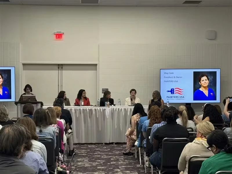 Conference room panel discussion with several speakers seated at a long table facing an audience; two large screens display a headshot and the text “Meg Cook, President & Owner, Painters USA.”