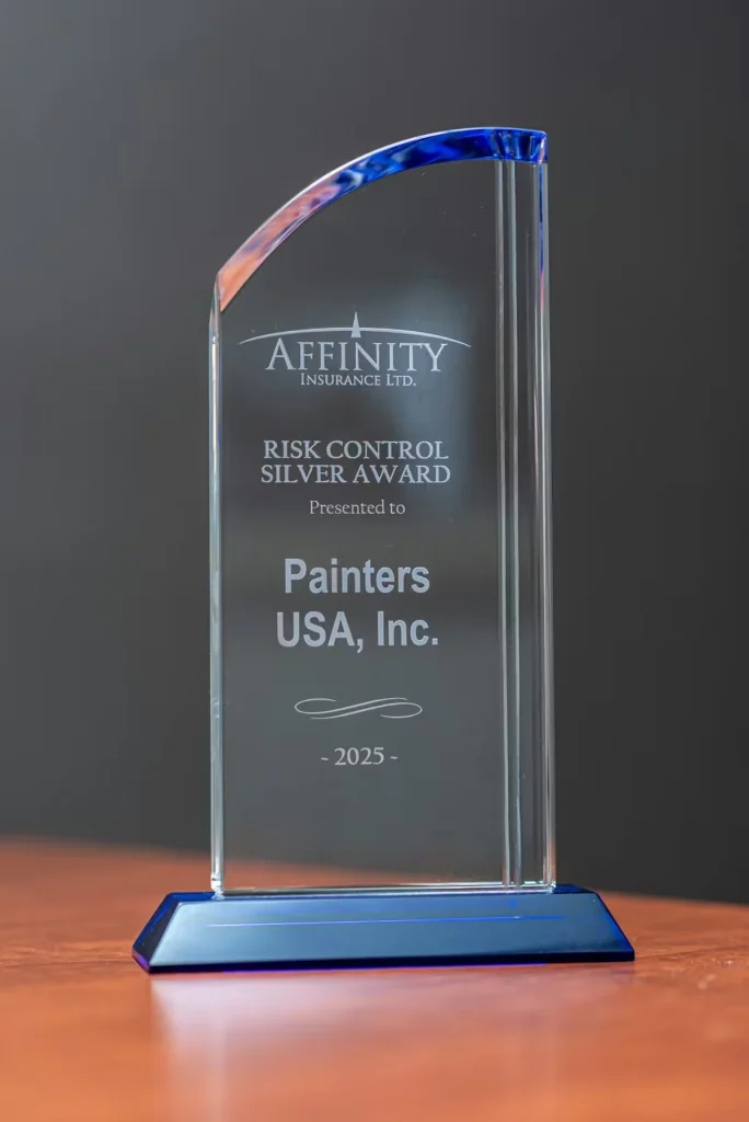 Clear glass award with a blue base on a wooden surface, engraved with “Affinity Insurance Ltd.” and “Risk Control Silver Award,” presented to “Painters USA, Inc.” with the year “2025.”