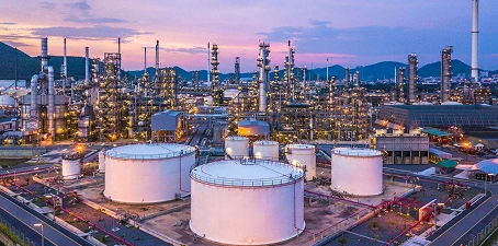 Wide view of a chemical plant at dusk with multiple storage tanks, towers, and pipelines brightly lit against the evening sky.