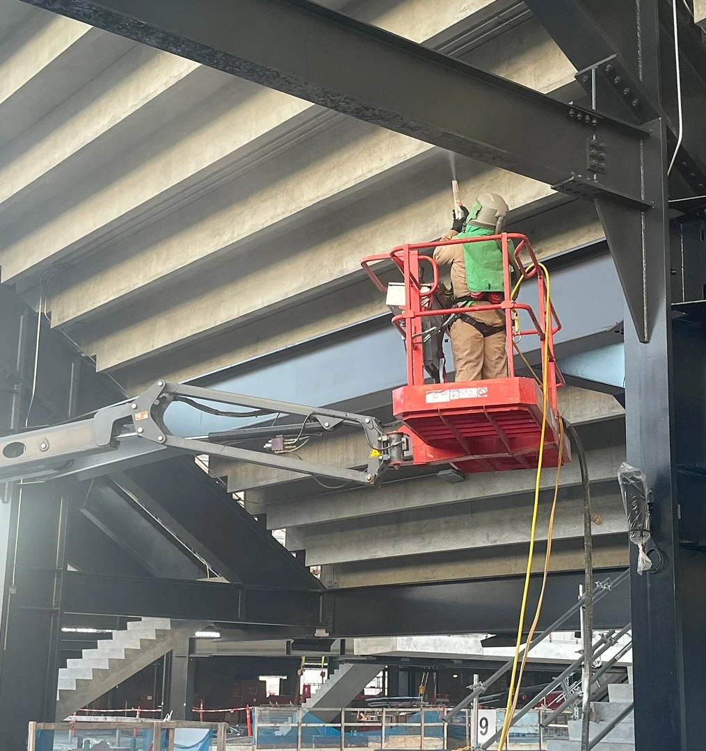A worker in protective gear operates a lift to work on structural steel beams, standing above a newly coated high-quality floor in a large industrial space.