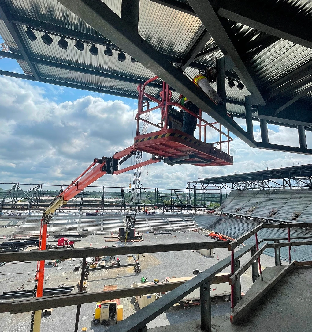 A construction worker in a safety harness operates a cherry picker lift, working on steel beams under a partially roofed stadium structure with open sky in the background.