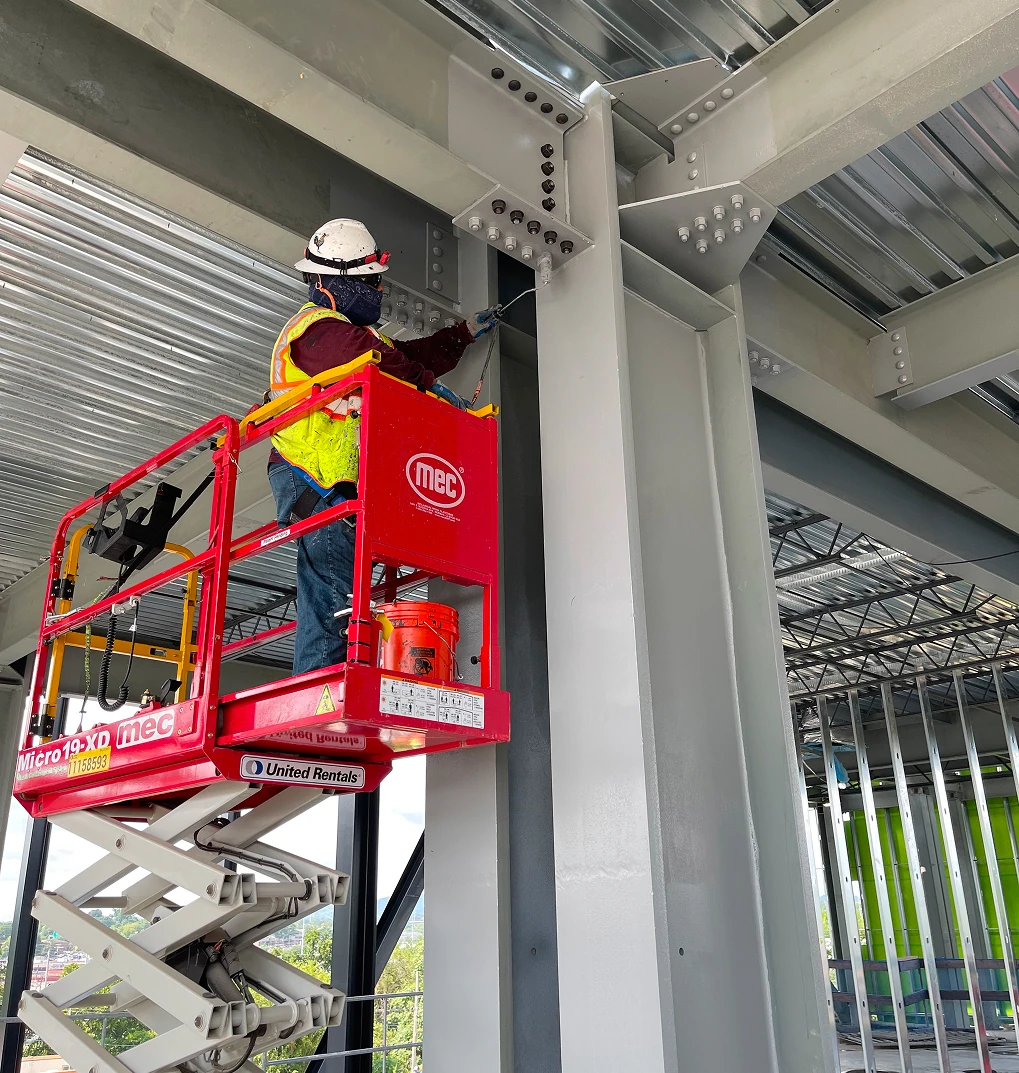A worker in protective gear and a green safety vest uses a red lift to reach and work on large steel beams inside a stadium under construction.