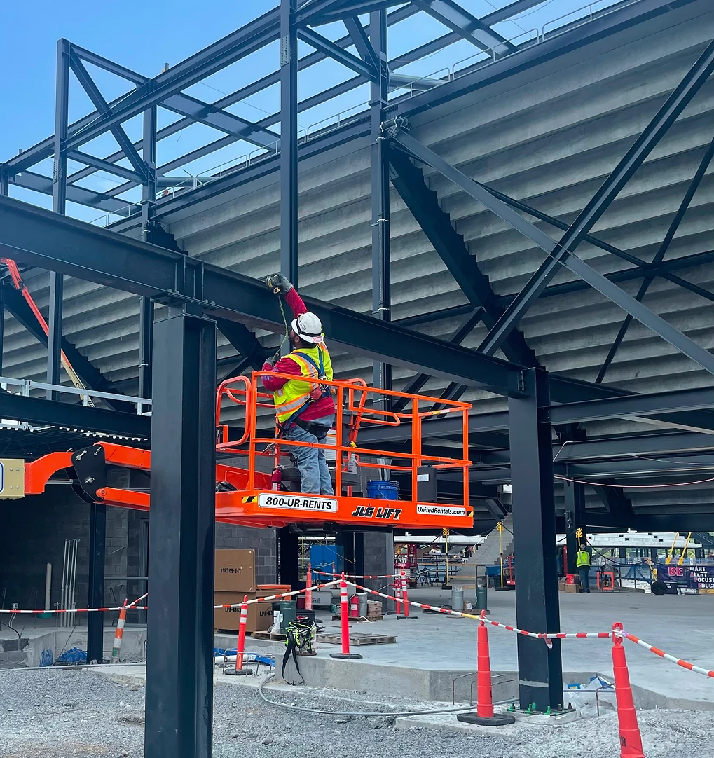 A construction worker in a hard hat and reflective vest stands on an orange lift, working on steel framework at an outdoor stadium construction site.