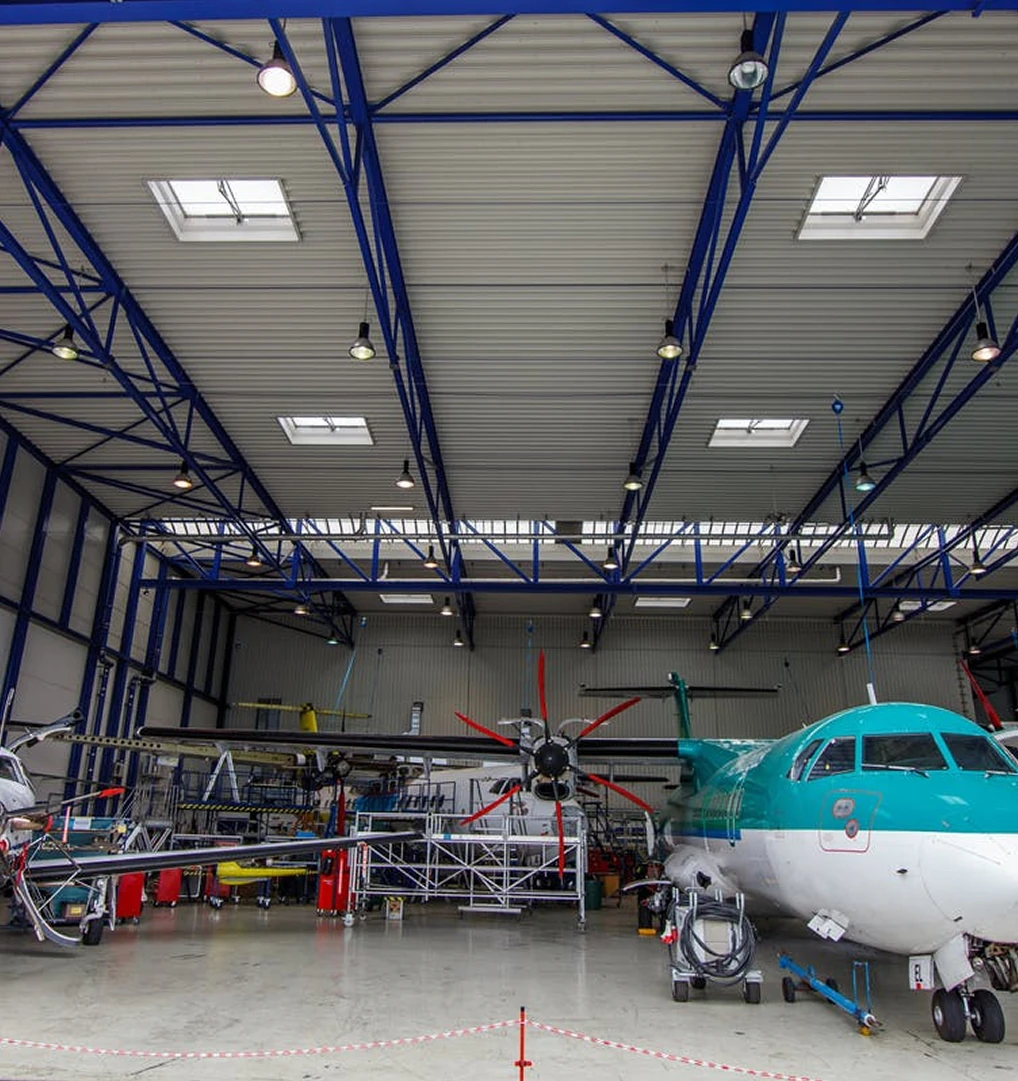 Interior of an aircraft hangar with a teal and white airplane under maintenance, surrounded by scaffolding and equipment beneath a high ceiling with skylights.