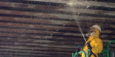 Power washing—worker in protective gear spraying high-pressure water on an overhead ceiling surface.