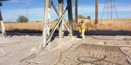 Sponge blasting—worker blasting paint or residue off outdoor steel beams with containment sheets on the ground.