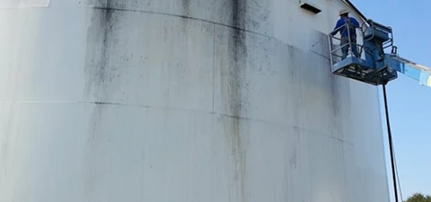 Worker on a lift applying protective coating to the side of a large white industrial storage tank with visible staining.