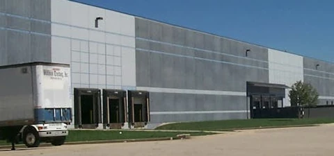 Exterior of a large gray warehouse with multiple loading docks, a semi-truck parked nearby, and clear blue sky above.