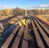 Worker in protective gear performing abrasive blasting on large steel beams outdoors, preparing surfaces for coating or restoration.