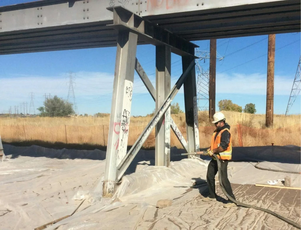 Worker in hard hat and orange vest abrasive-blasting graffiti-marked steel bridge columns over plastic containment sheeting in an open field.