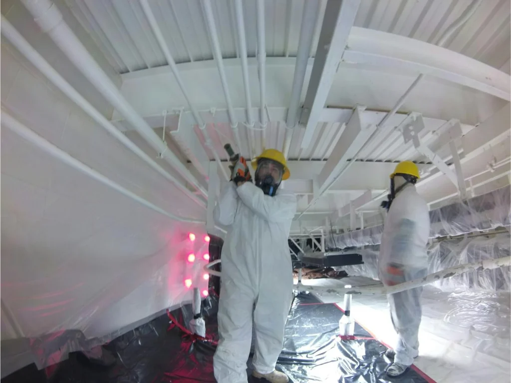 Two technicians in Tyvek suits and respirators preparing or coating overhead pipes in a plastic-contained room with curing lights on.