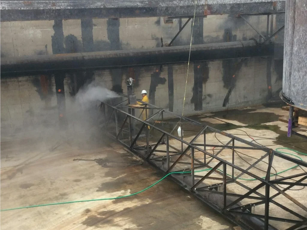 Worker in yellow PPE abrasive-blasting a large steel truss inside an industrial pit; dust and mist drift across the concrete floor.