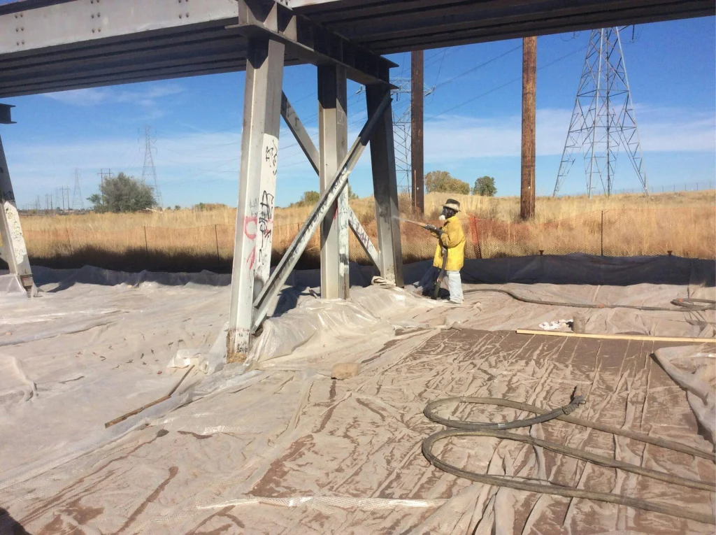 Two workers blasting steel support columns under a bridge; plastic sheeting covers the ground for containment in a grassy right-of-way.