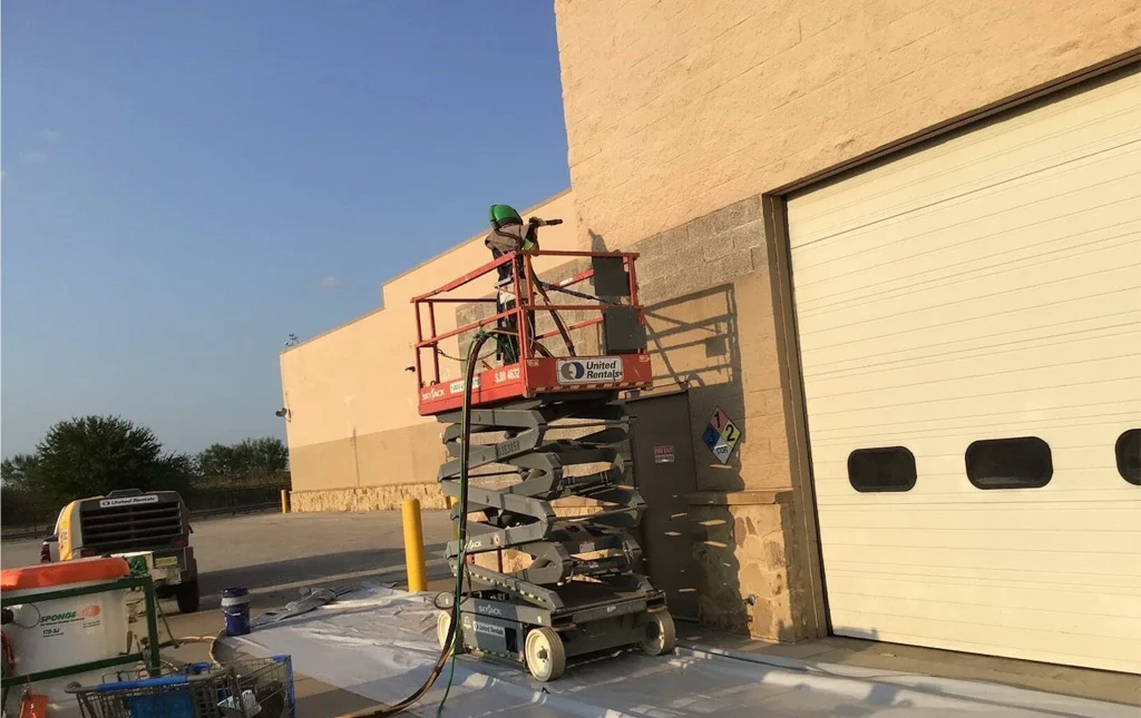 Operator on a scissor lift blasting the exterior CMU wall of a warehouse near a roll-up door, with ground tarps and hose lines below.