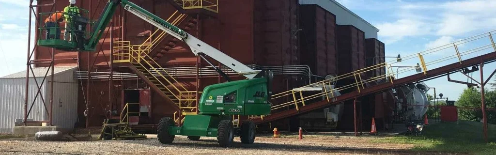 Two workers on a green boom lift servicing a large industrial building with yellow stairs and an angled conveyor.