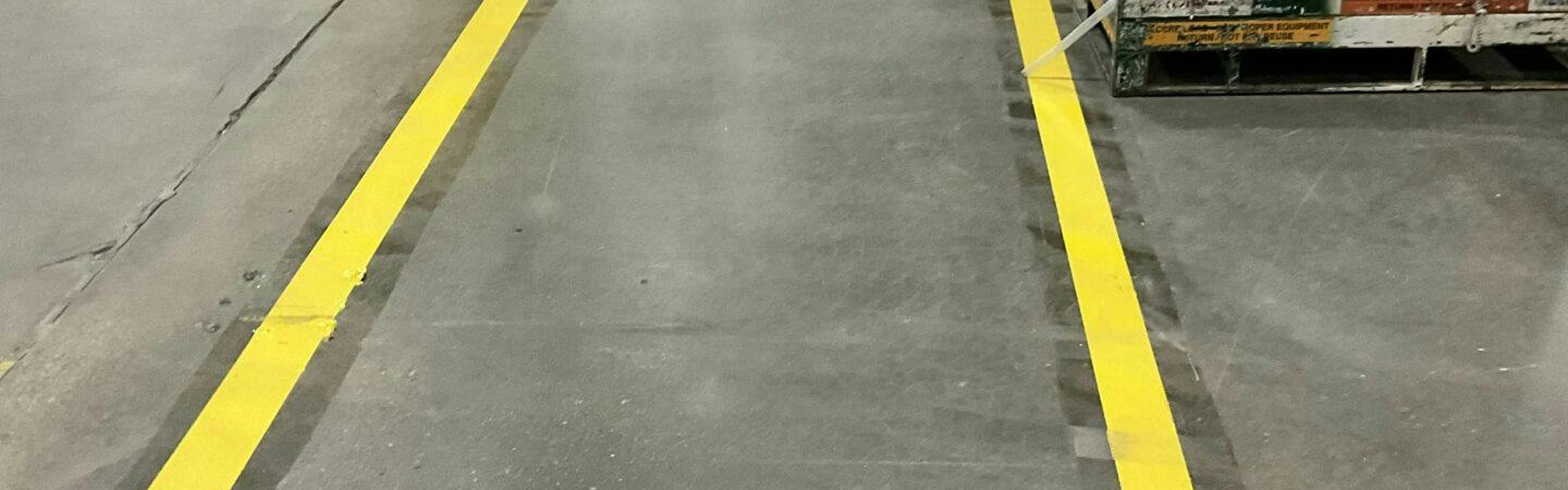 Two parallel yellow safety lines marking a straight aisle on warehouse concrete.