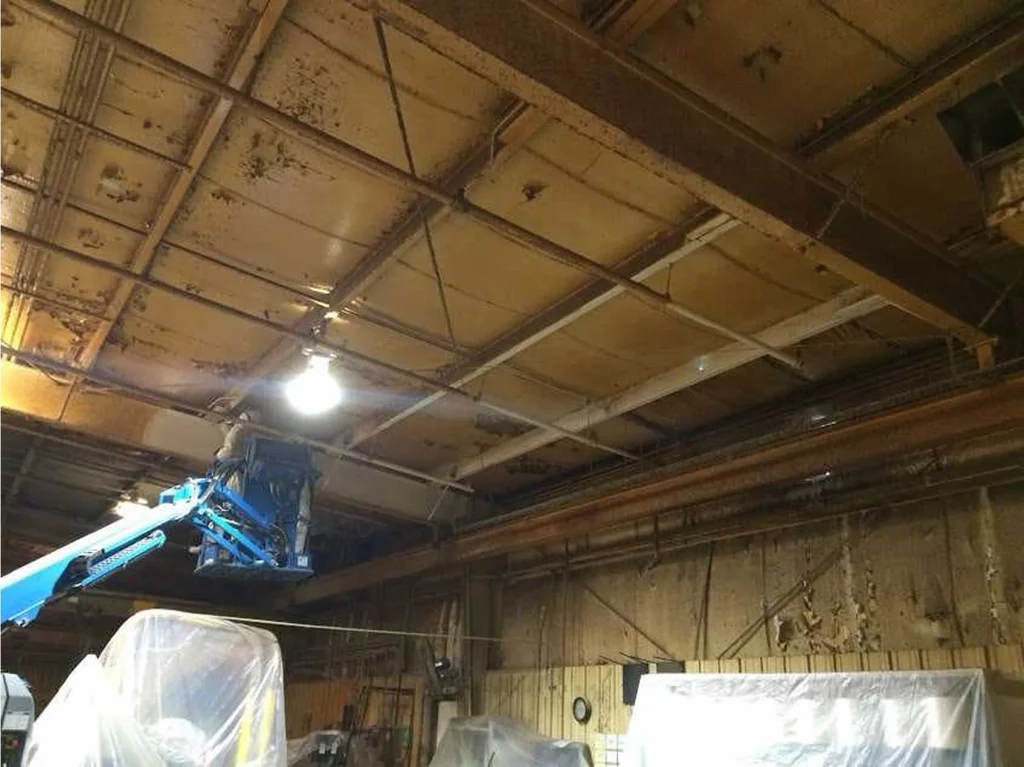 Industrial shop before cleaning, showing a heavily stained metal ceiling and beams with rust and residue; a blue boom lift is raised toward the ceiling above plastic-covered equipment.